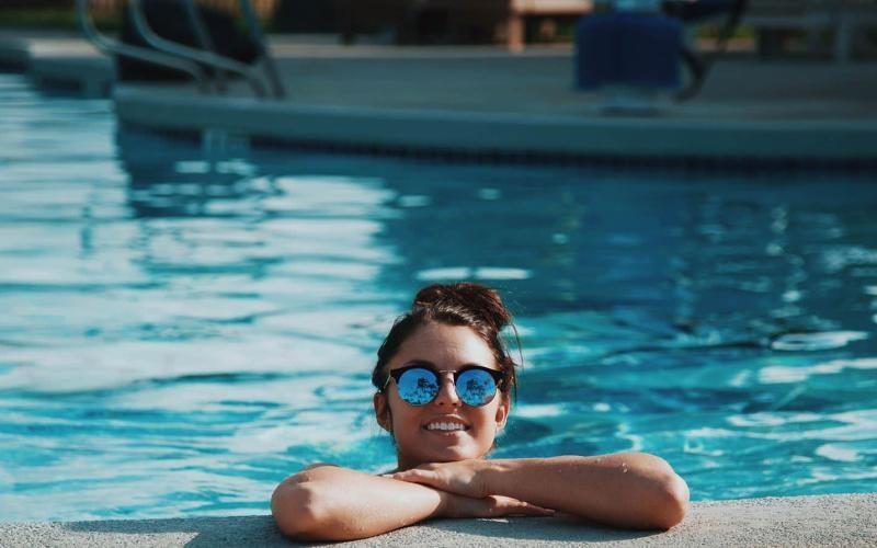 woman with sunglasses, in a pool, rests her arms on the ledge and smiles